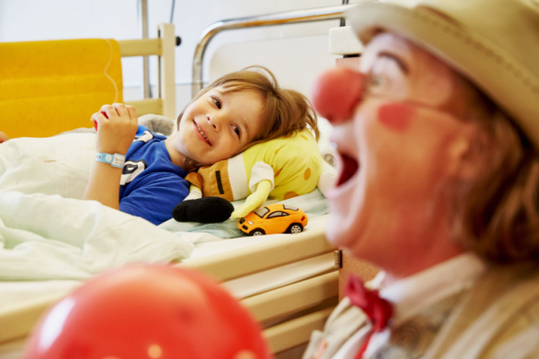 Ein lachendes Kind liegt im Krankenhausbett mit Plüschtier und gelbem Kissen, vorne ein Clown mit roter Nase