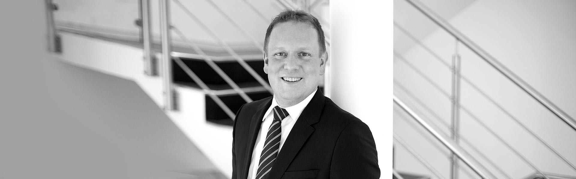 Ein lächelnder Mann im Anzug vor einer Treppe. Image in Schwarz-Weiß mit einem freundlichen Ausdruck.