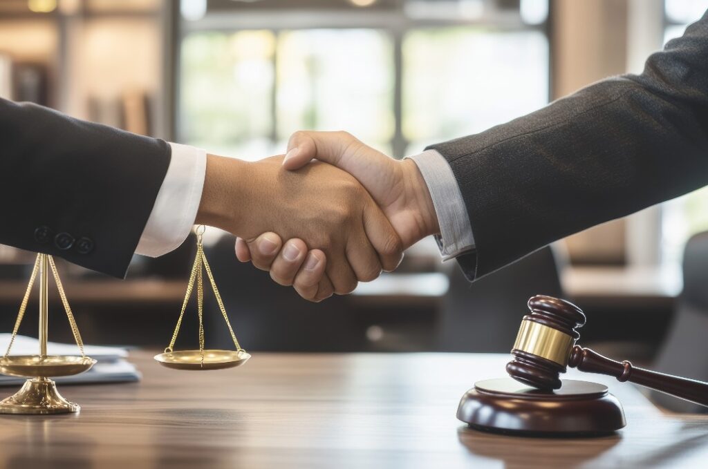 Zwei Anwälte schütteln sich die Hände in einem modernen Büro, mit einer Waage und einem Hammer auf dem Schreibtisch. Dies symbolisiert ihre Einigkeit und Partnerschaft.